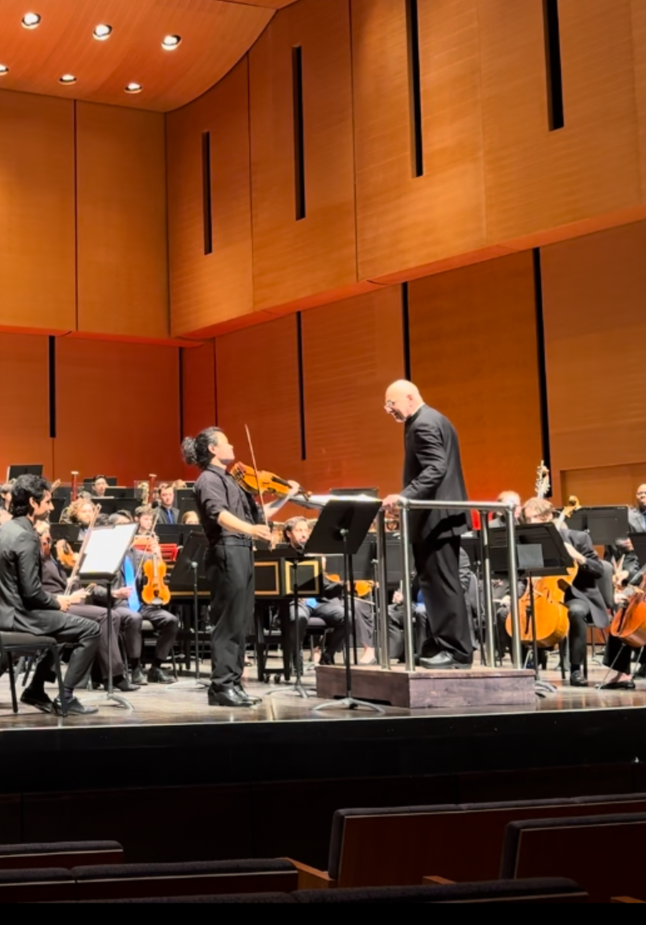 A soloist standing beside a conductor, with an orchestra behind them.